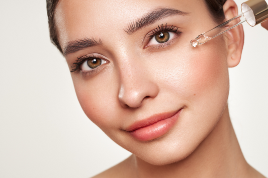 Applying skincare: A close-up of a woman's face as she holds a dropper bottle, dispensing face oil onto her cheek. Image for blog post:5 Reasons You Should Be Using a Face Oil