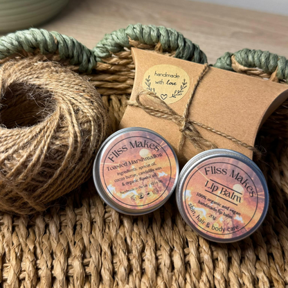 Two tins of 100% natural handcrafted toasted marshmallow lip balm made with organic, vegan and cruelty-free ingredients, showcasing its smooth texture. In the background, there are nature loving accessories, emphasising the product's organic composition. 